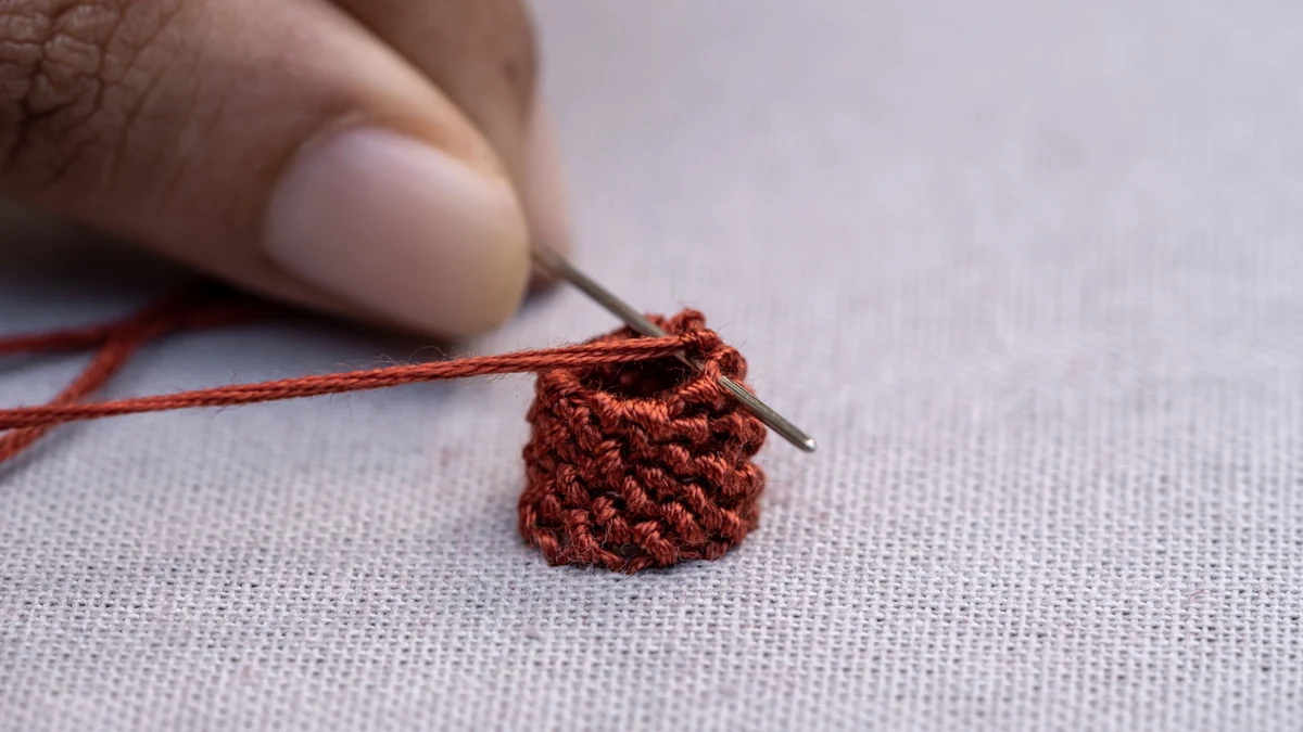 A needle working reddish-brown thread into a raised detail on pale fabric.